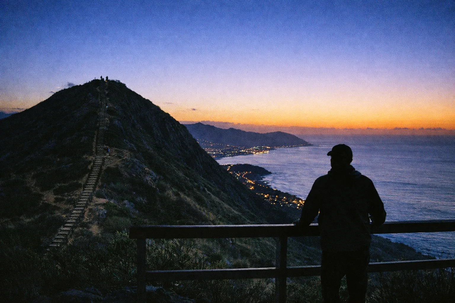 Person standing at a railing overlooking a mountain path, ocean, and coastline at sunset