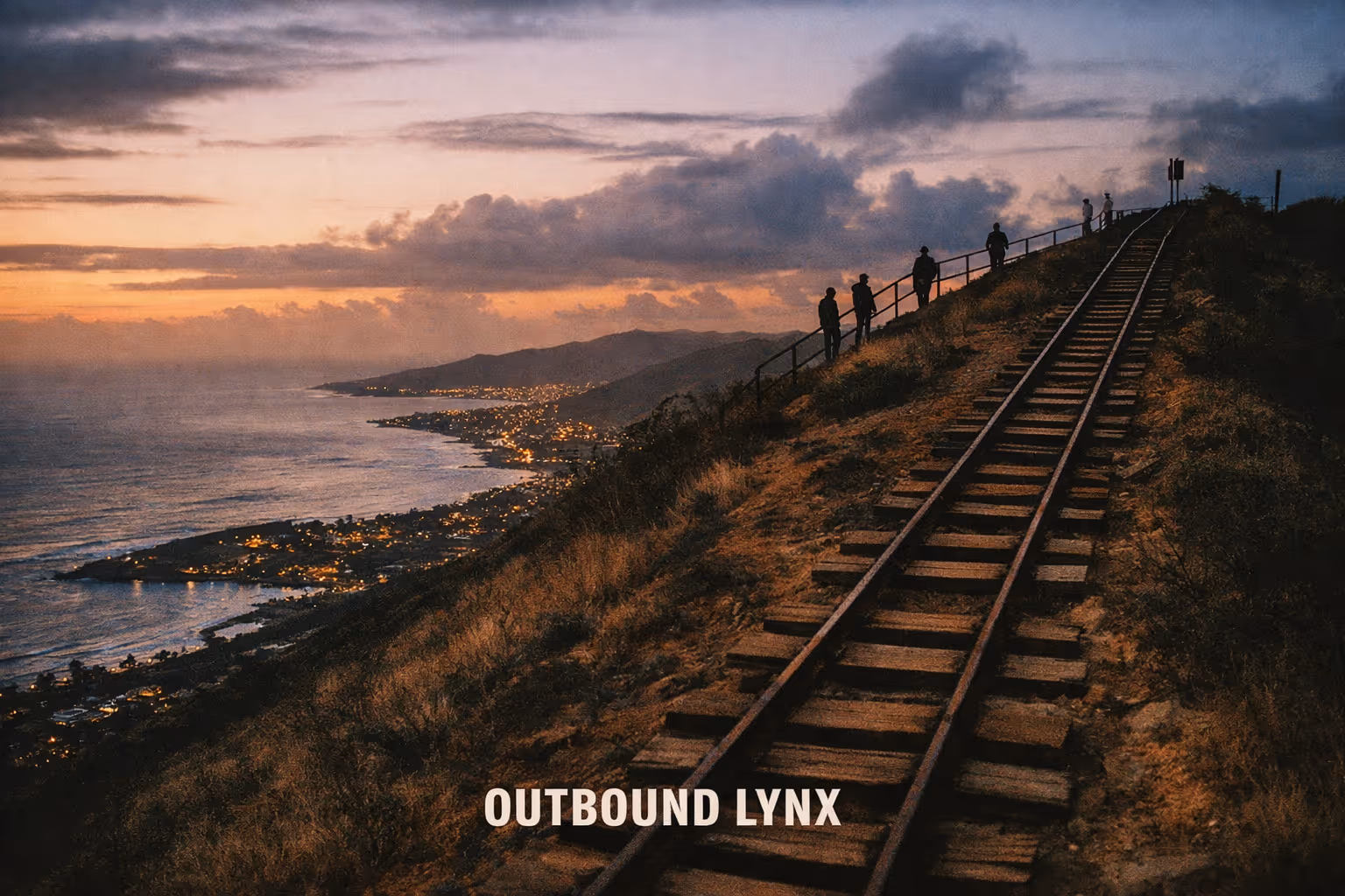 Railway tracks on a hillside at sunset with several people standing by a fence overlooking a coastal city