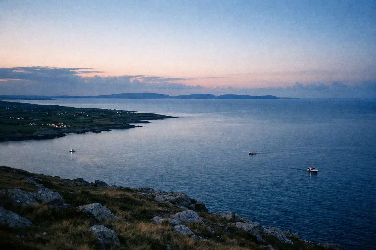 Aran Islands sit off the Irish coast, seen from a rocky shoreline with boats in the water at dusk.