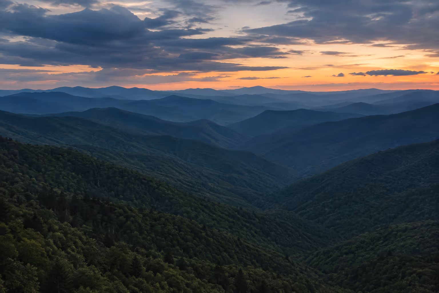 Layered forested ridges of the Great Smoky Mountains stretch beneath a sunset sky.