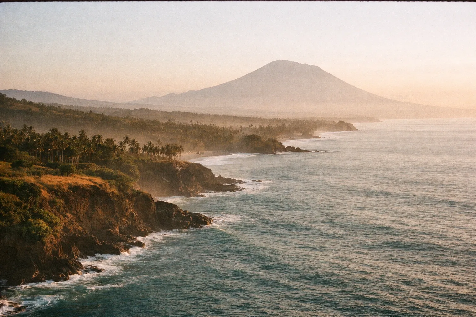Aerial view of Bali’s coastline at sunrise with Mount Agung in the distance and palm trees lining the shore