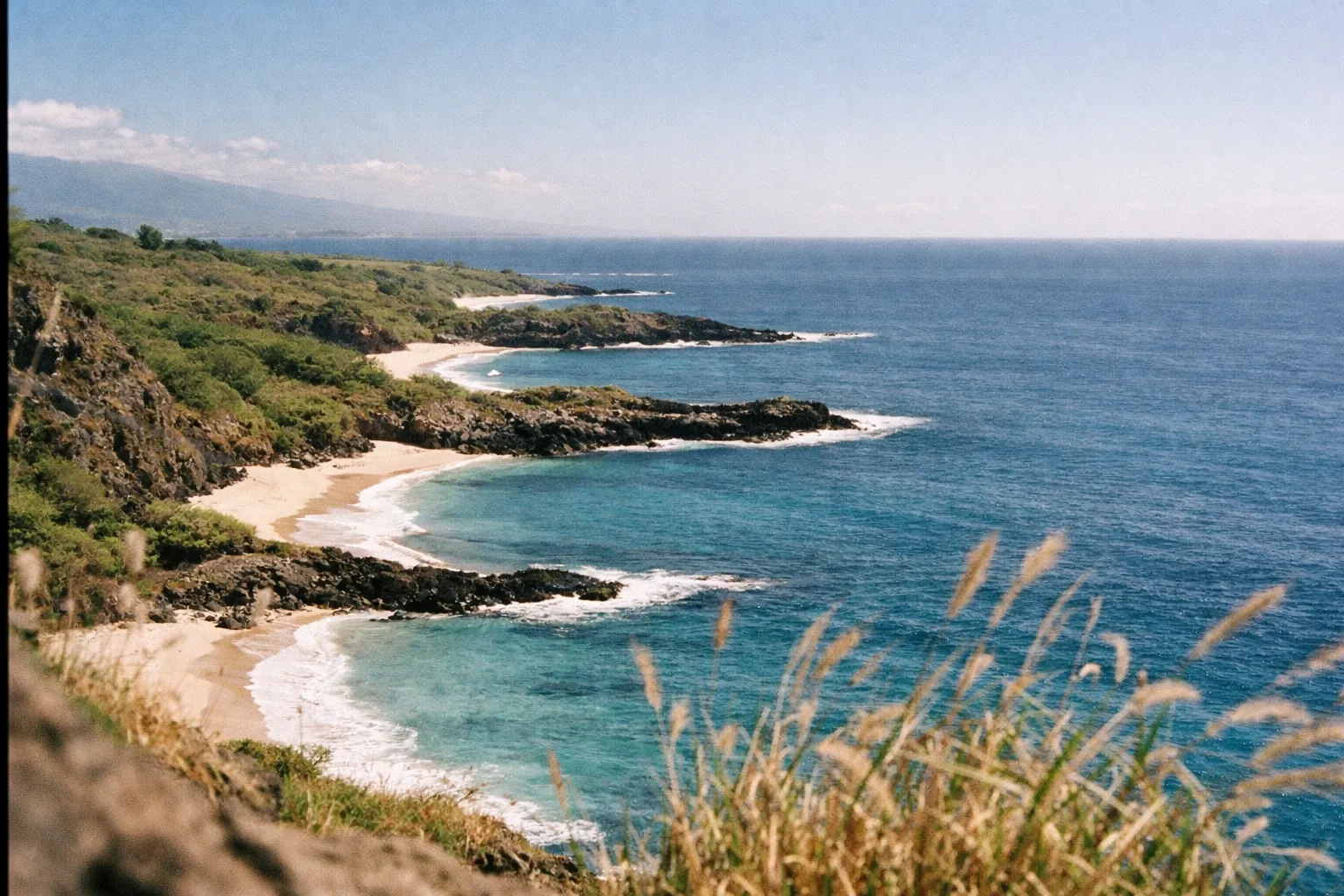 A series of secluded sandy coves line a rugged coastline beside clear blue ocean under a sunny sky.