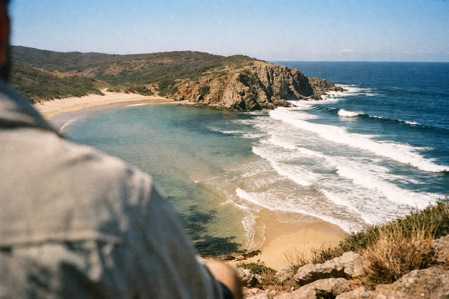 A person sits on a cliff overlooking a sandy beach and rocky coastline with waves rolling in.