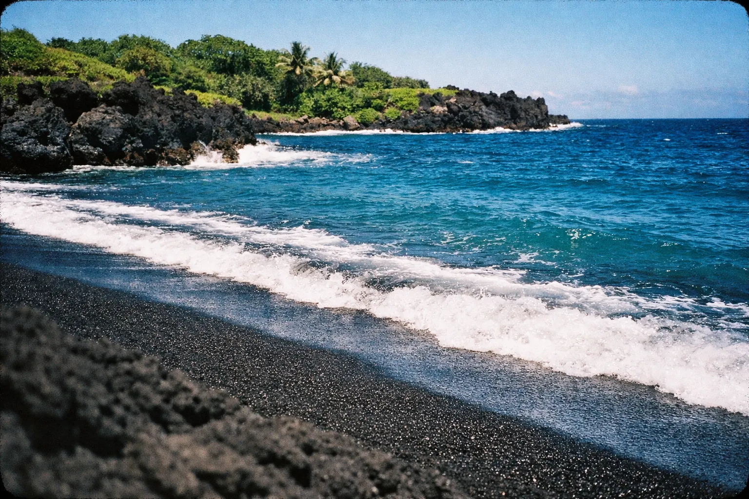 Waves wash onto the black sand and rocky shore at Waianapanapa State Park, Maui, with lush greenery in the background.