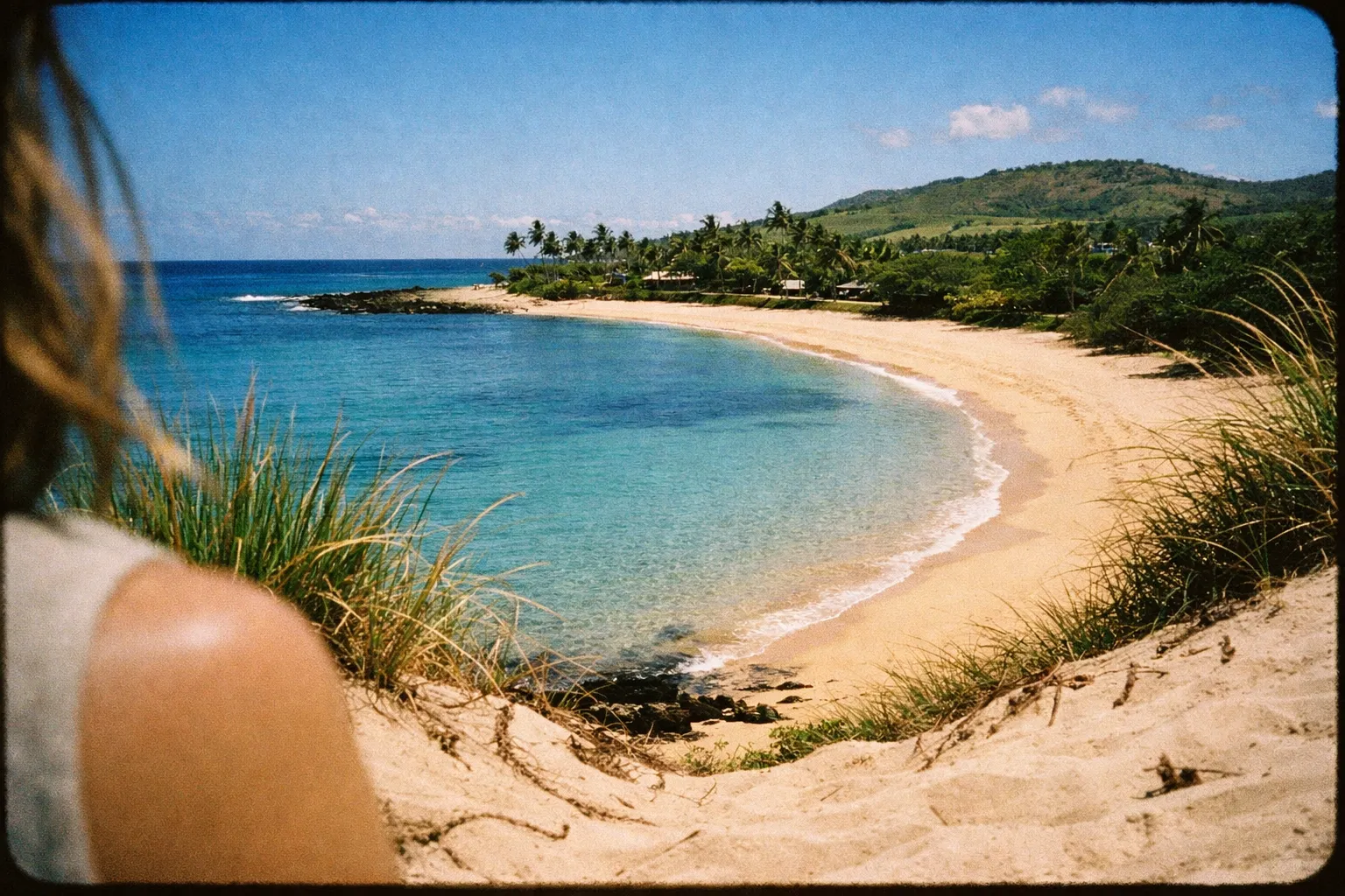 A person sits on a sandy dune overlooking a tropical beach with clear blue water and palm trees.