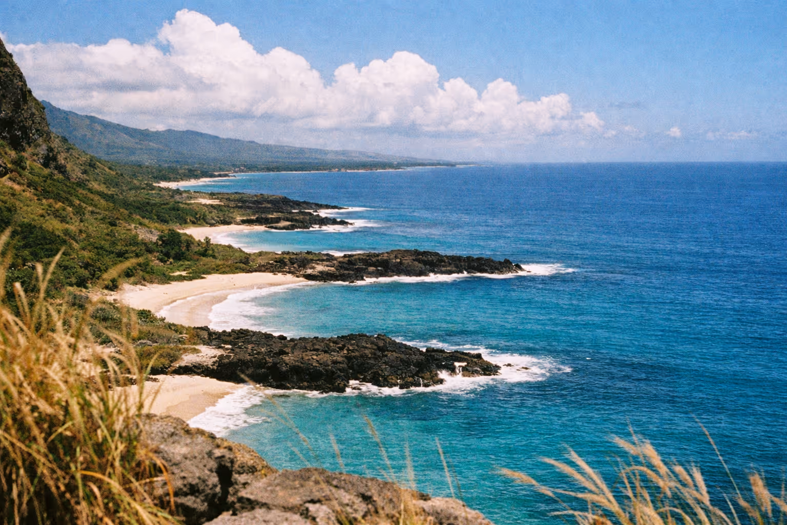 A view of a rugged coastline with sandy beaches and turquoise water backed by green hills in Hawaii.