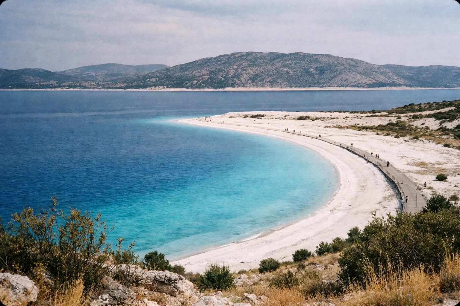 People walk along the curved white sand shore of Salda Lake beach with mountains in the background.
