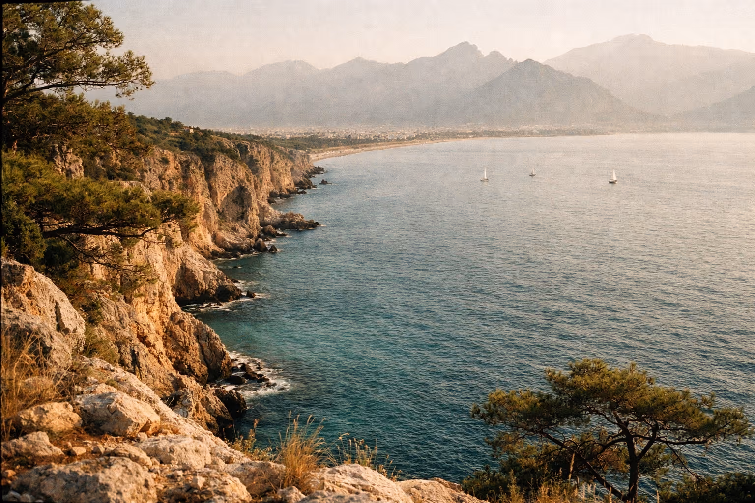 Rocky cliffs and pine trees overlooking the Mediterranean Sea near Antalya, with mountains in the distance.