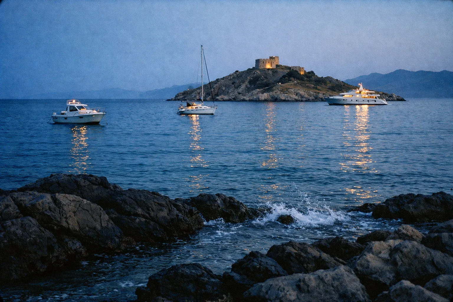 Yachts anchored near a rocky island with a stone fort at dusk, viewed from the shoreline.