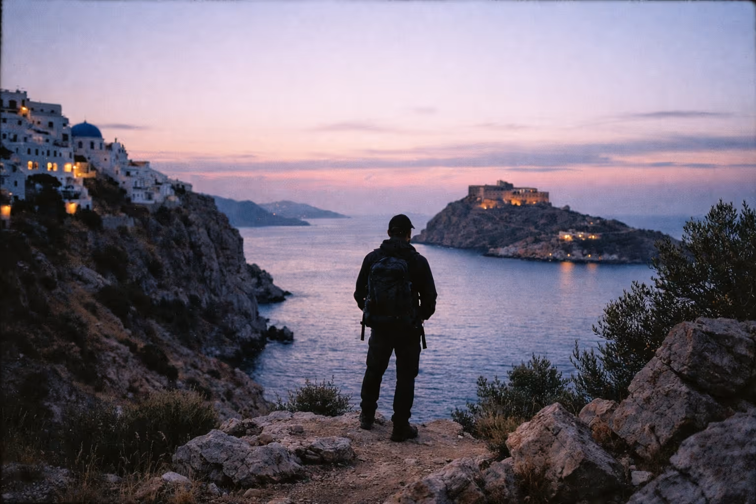 A person with a backpack stands on a cliff overlooking white buildings and a domed church by the sea at sunset.