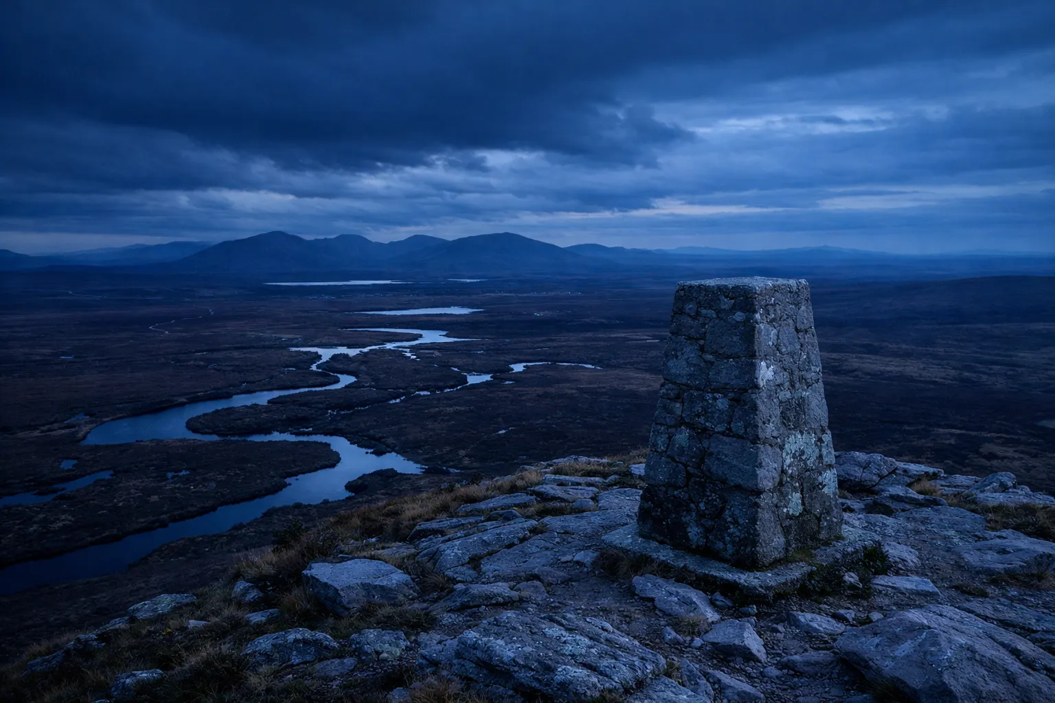 A stone marker stands on a rocky hill overlooking winding rivers and distant mountains in Connemara National Park.