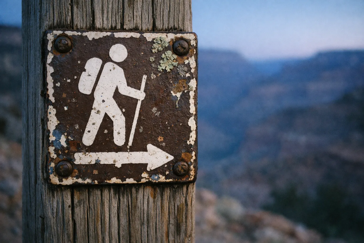 A weathered hiking trail sign is mounted on wood with a canyon landscape in the background.
