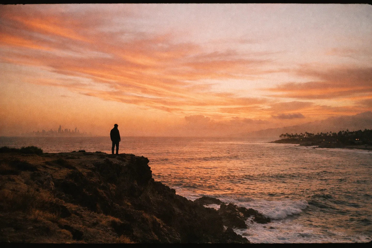 Silhouette on a cliff at golden hour with distant NYC skyline on the left and Maui coastline on the horizon.
