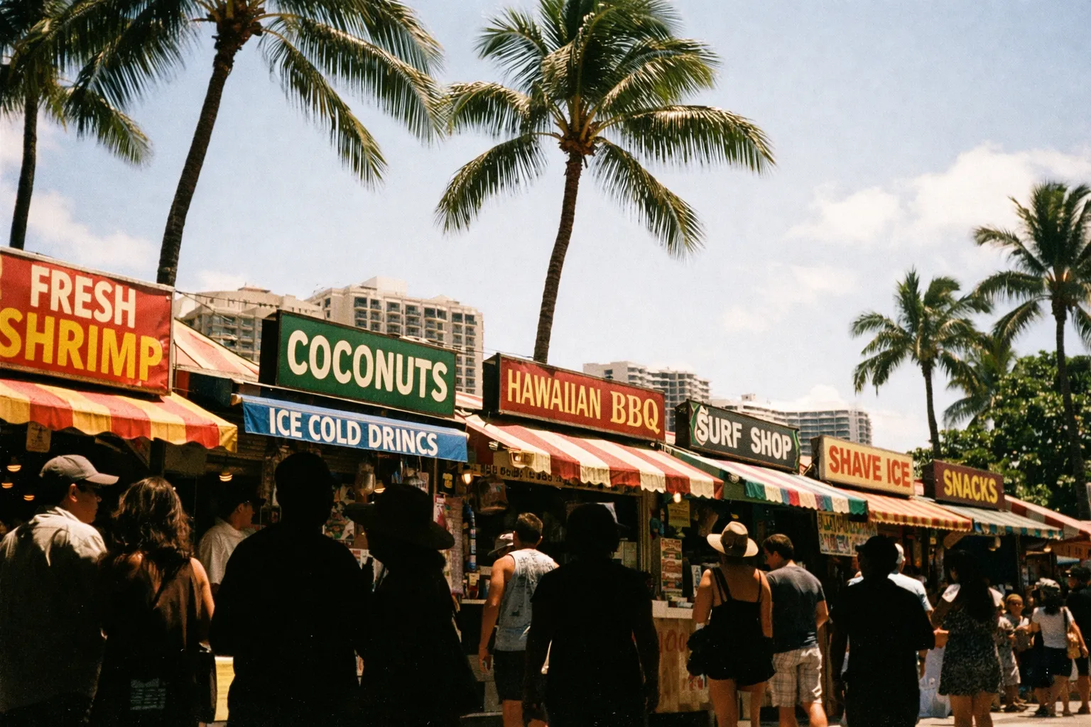 People browse outdoor food and surf stalls under palm trees, with high-rise hotels in the background in Waikiki, Oahu.