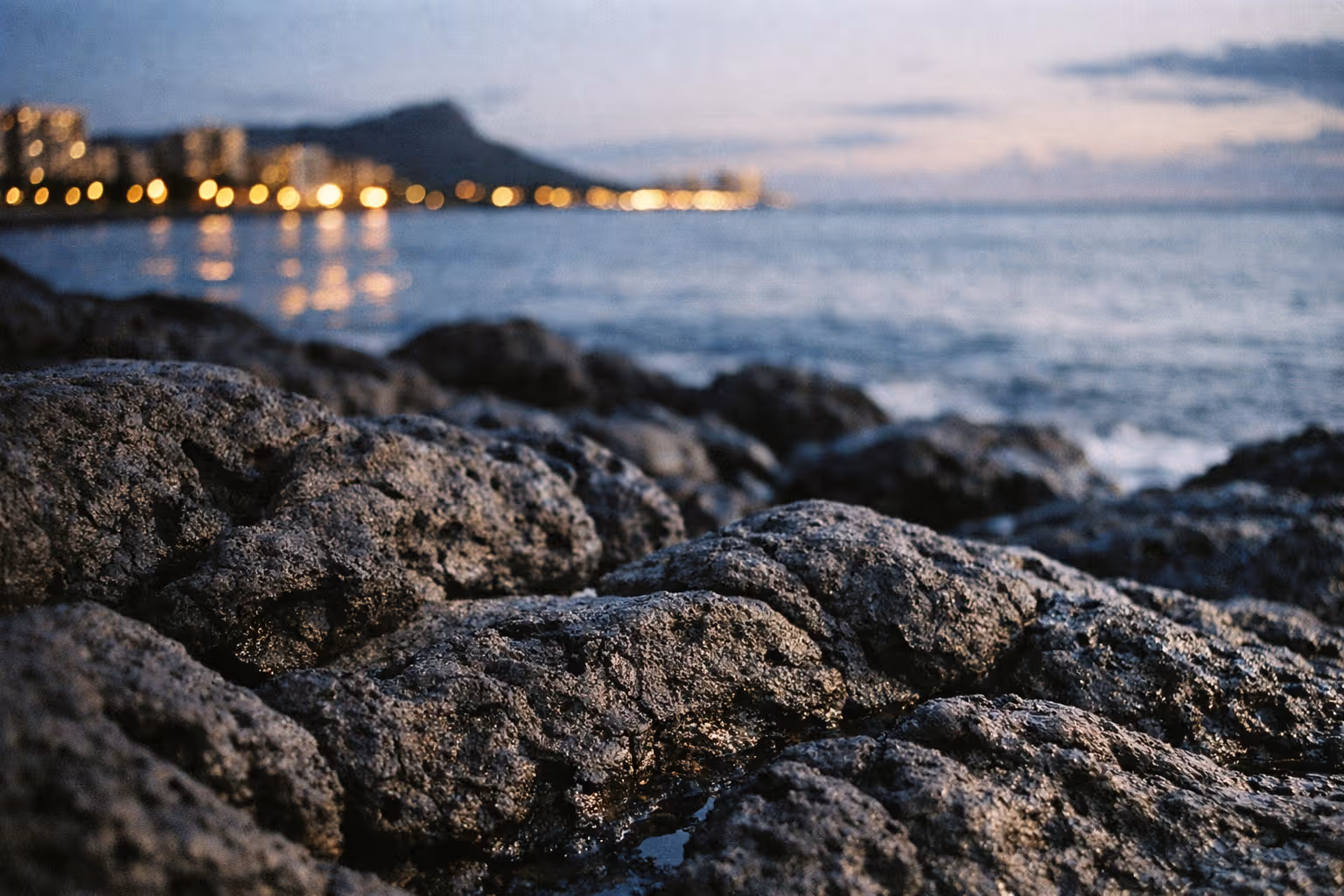 Lava rocks on the shore at sunset with Diamond Head and Waikiki skyline in the distance.