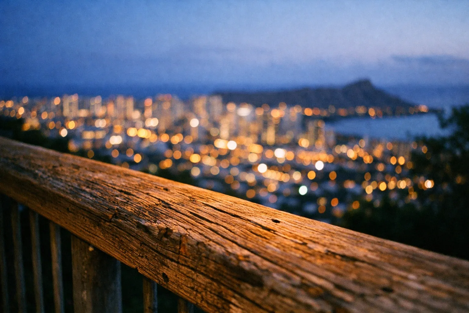 A wooden railing overlooks Honolulu city lights and Diamond Head at dusk from an elevated viewpoint.