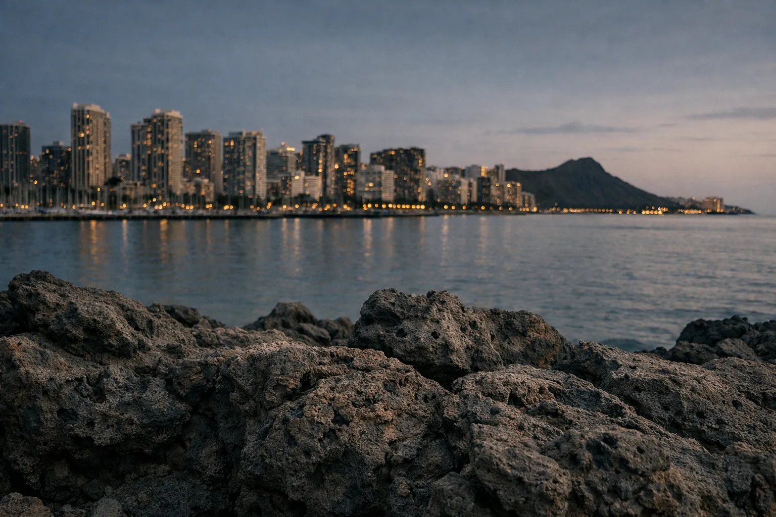 Rocky shoreline with a city skyline and Diamond Head mountain in the background at dusk.