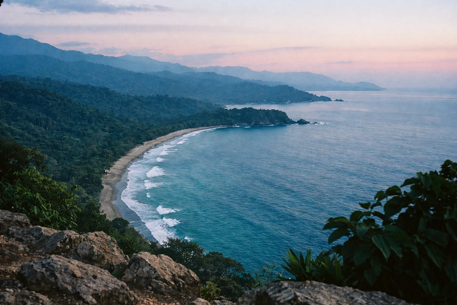 Dense rainforest meets a curving coastline and beach in a tropical landscape at dusk.