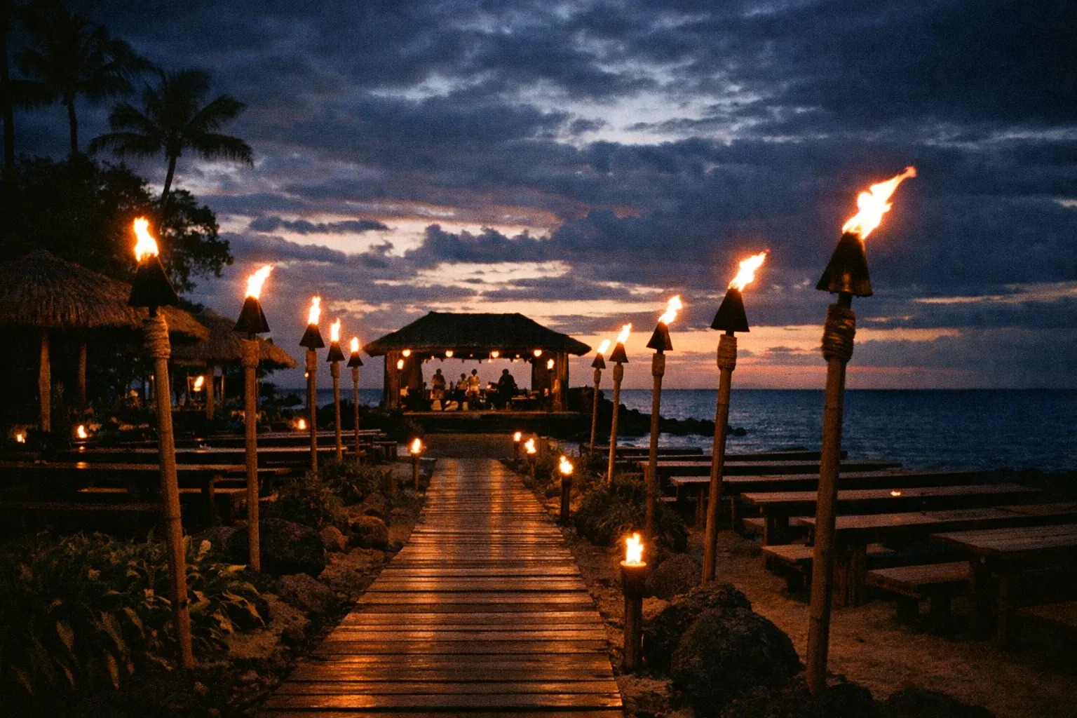 Evening luau venue on Hawaii island with tiki torches and distant stage in silhouette