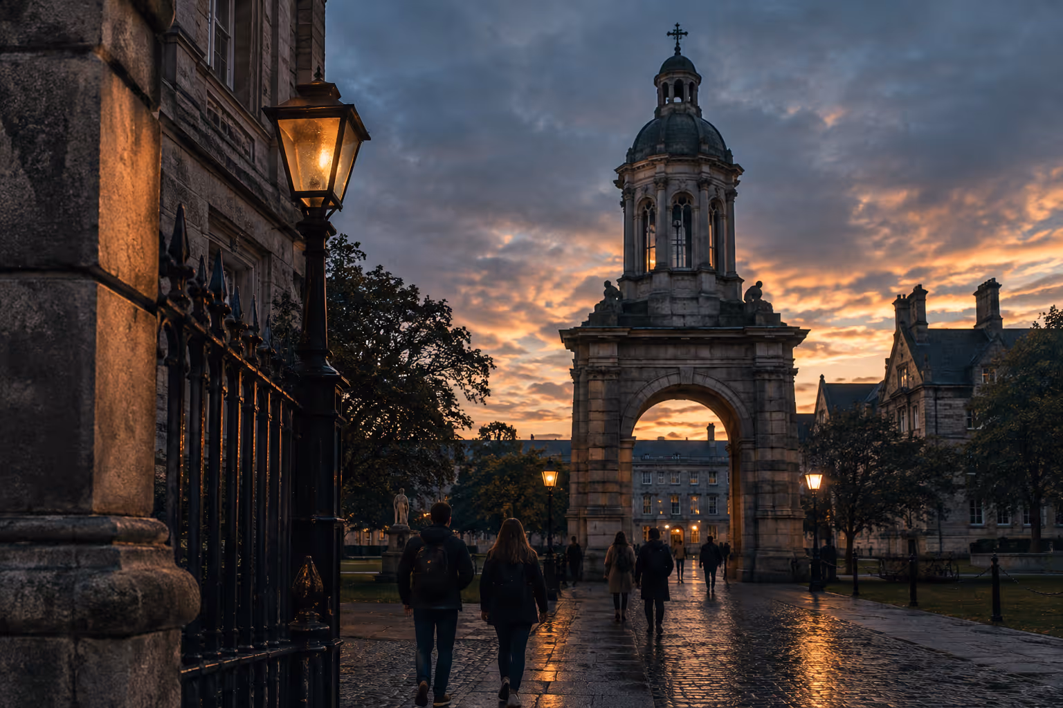 Visitors cross the cobbles of Parliament Square at Trinity College Dublin toward the Campanile at dusk.