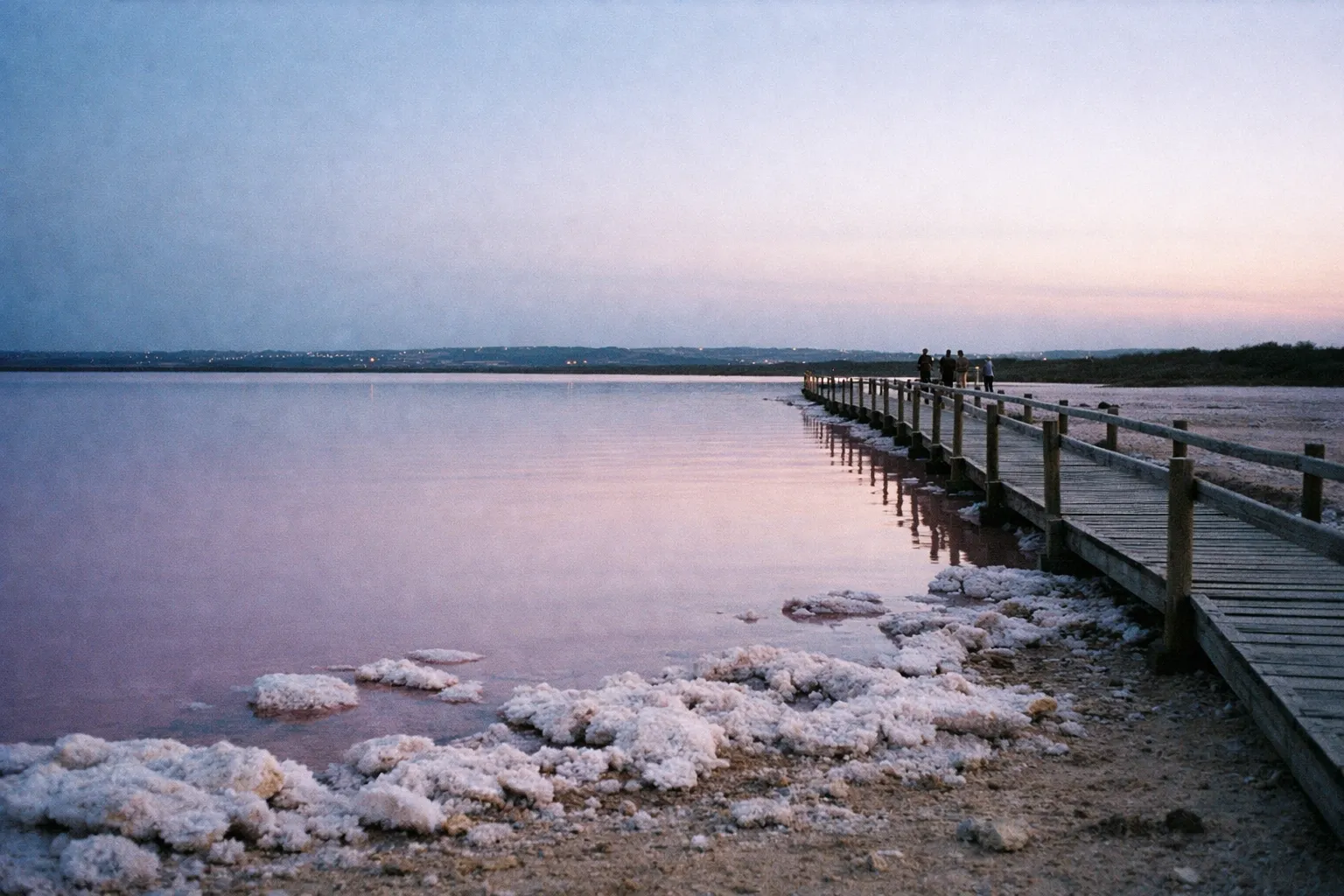A wooden walkway stretches over the pink salt flats of Cabo Rojo at sunset.