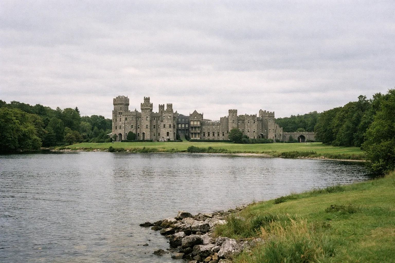 Ashford Castle overlooks a calm lake, surrounded by lush green lawns and trees in rural Ireland.
