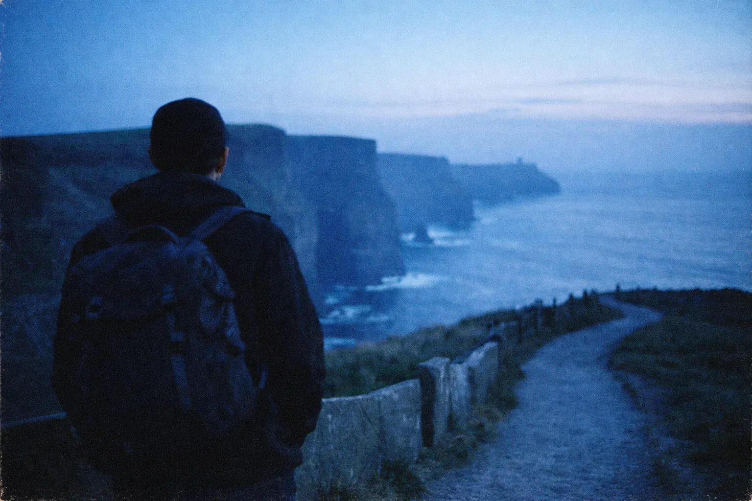 A person with a backpack stands on a trail overlooking the Cliffs of Moher at dusk.
