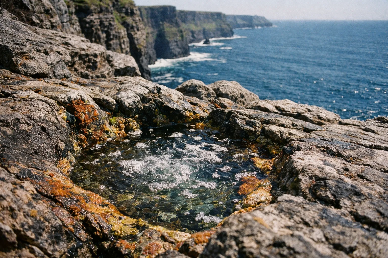 Rocky tide pool on a cliff edge with the Cliffs of Moher and the Atlantic Ocean in the background.