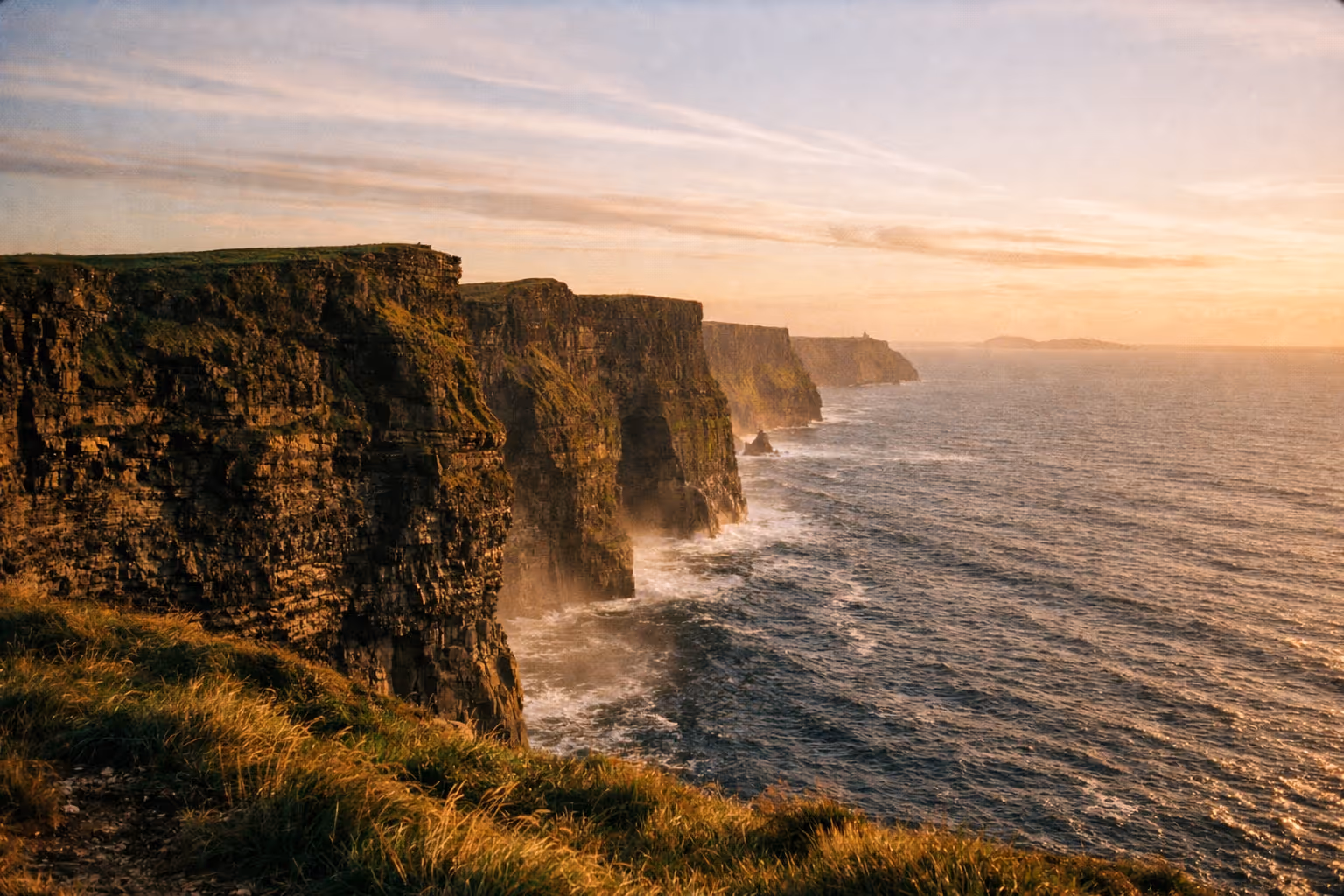 The Cliffs of Moher rise above the Atlantic Ocean at sunset with grassy edges in the foreground.