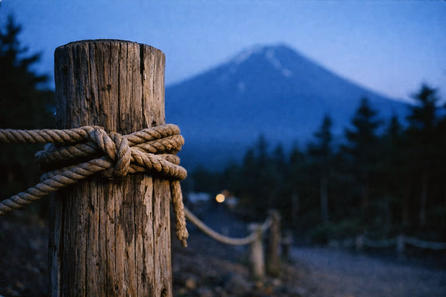 A wooden post with rope marks a trail leading toward Mount Fuji in the background at dusk.