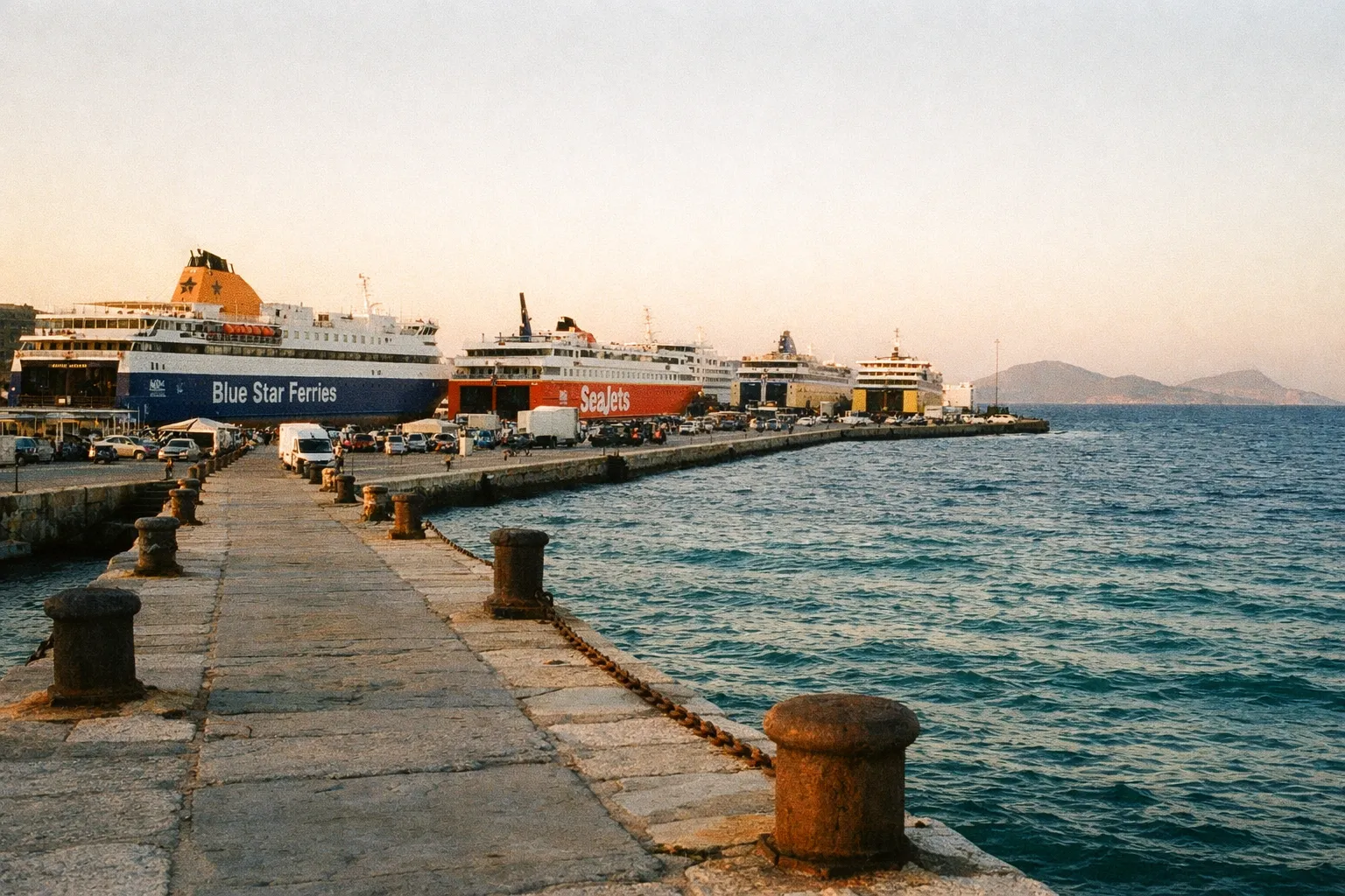 Ferries docked at a pier in a Greek island port with mountains visible across the water.