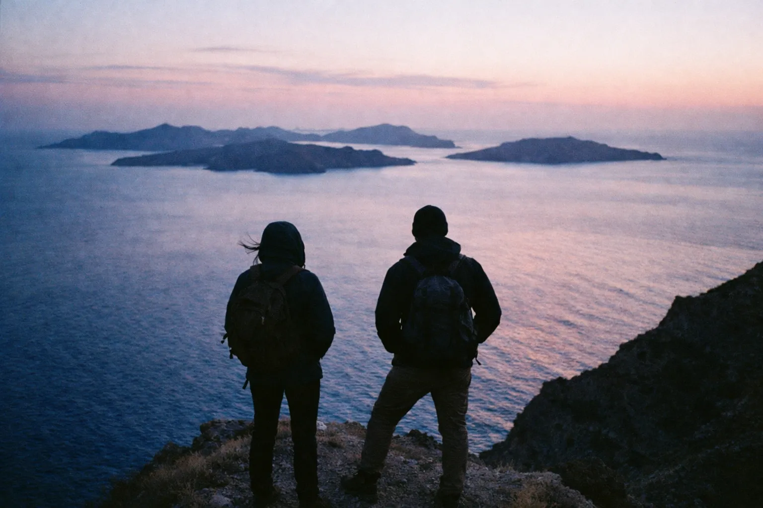 Two people with backpacks stand on a cliff overlooking several islands at sunset.