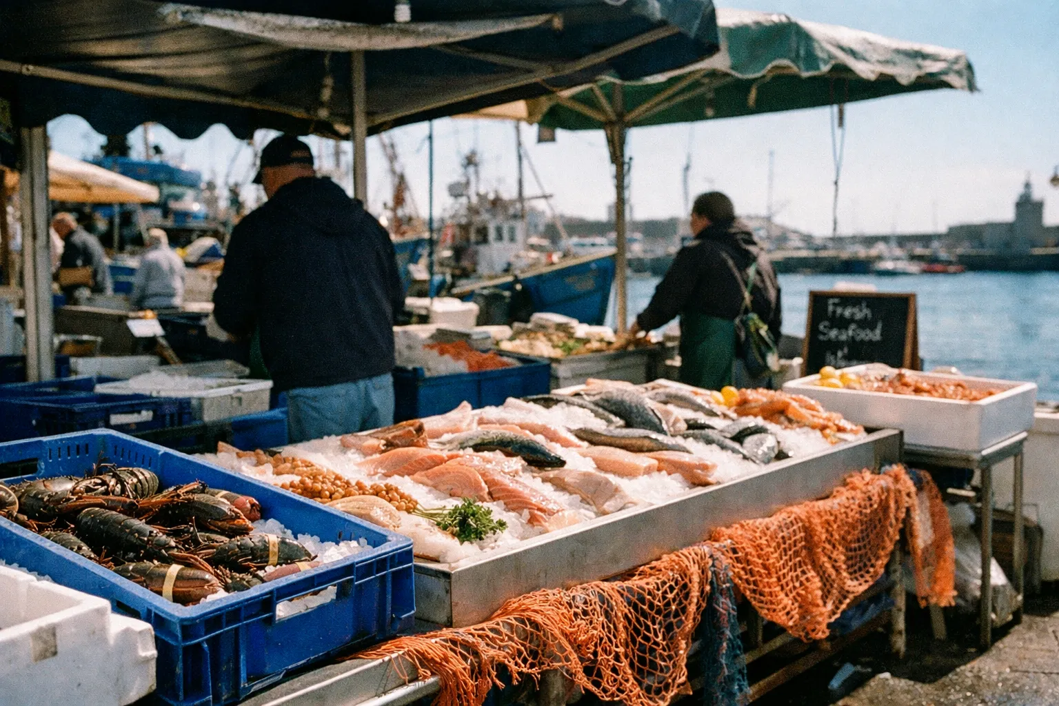 Fresh seafood displayed on ice at an outdoor market by the harbor with fishing boats in the background.