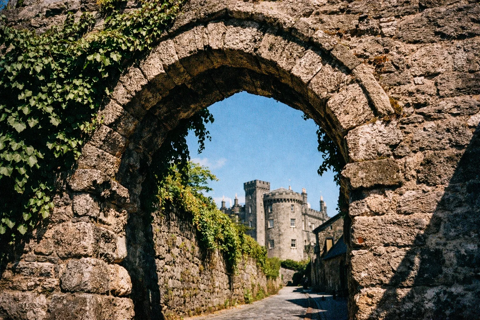 A stone archway frames a view of Kilkenny Castle under a clear blue sky.