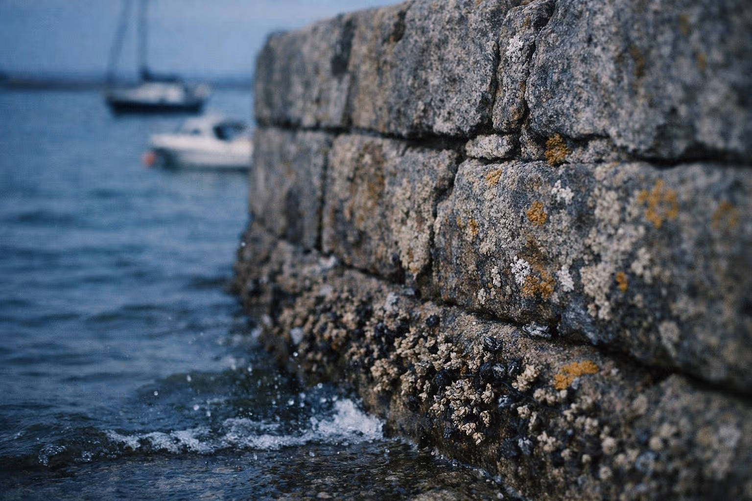 A close-up of a stone harbor wall with boats moored on the water in the background.