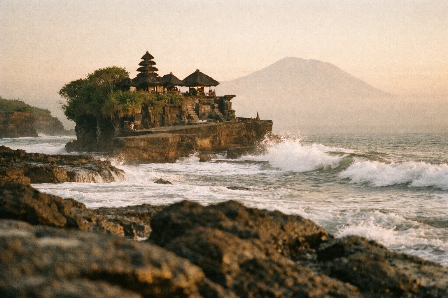 Balinese coastal temple at golden hour with a distant volcanic mountain.
