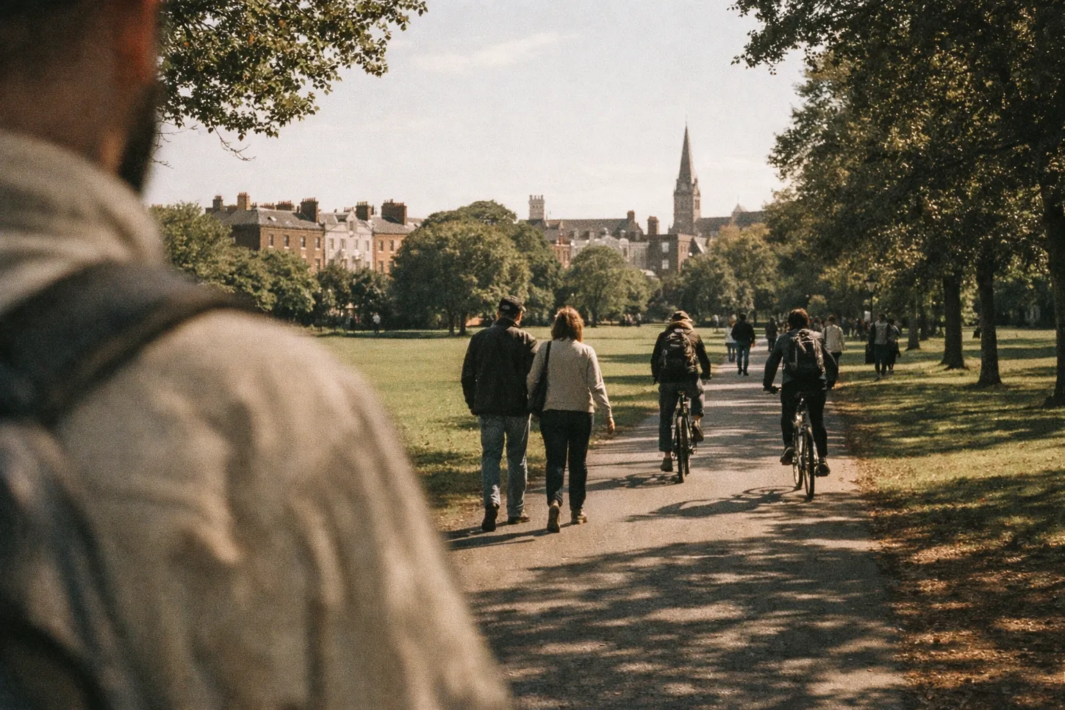 People walk and cycle along a tree-lined path in Dublin's St Stephen's Green with city buildings in the background.