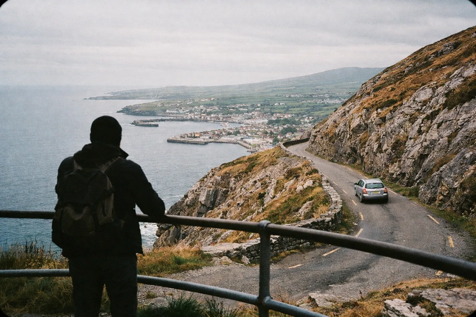 A person stands by a railing overlooking Dingle town and harbor from a winding coastal road along a rocky hillside.