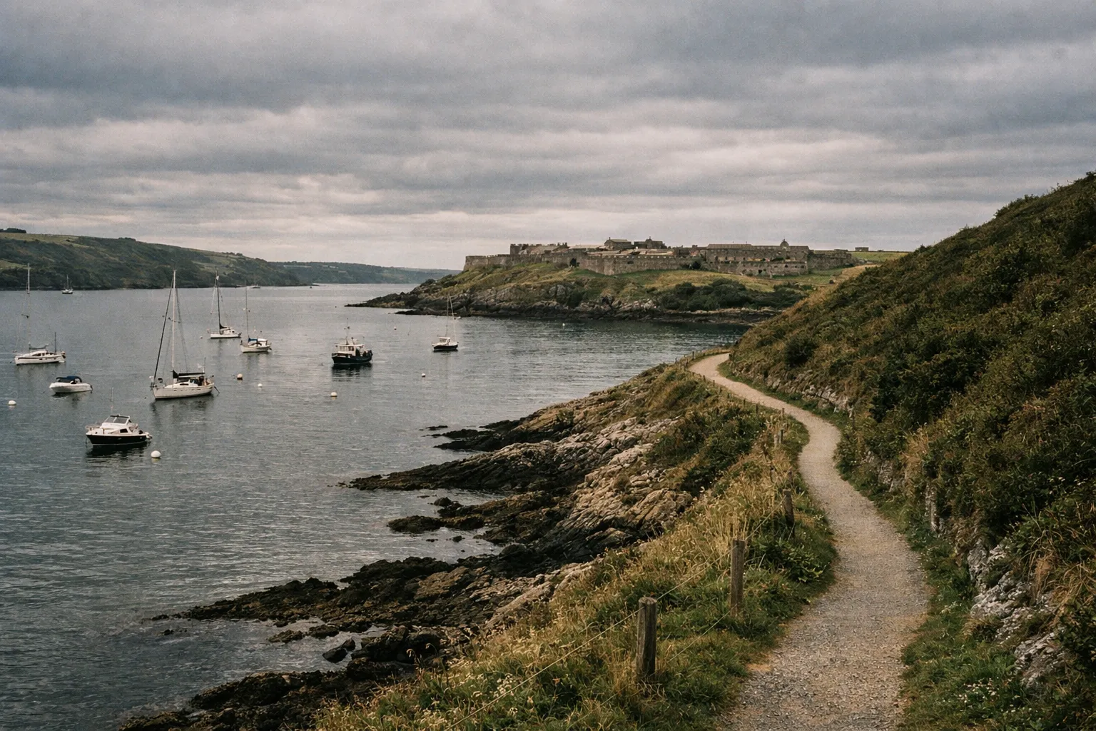 Sailboats anchored in a bay near Charles Fort and a winding coastal path in Kinsale, Ireland.
