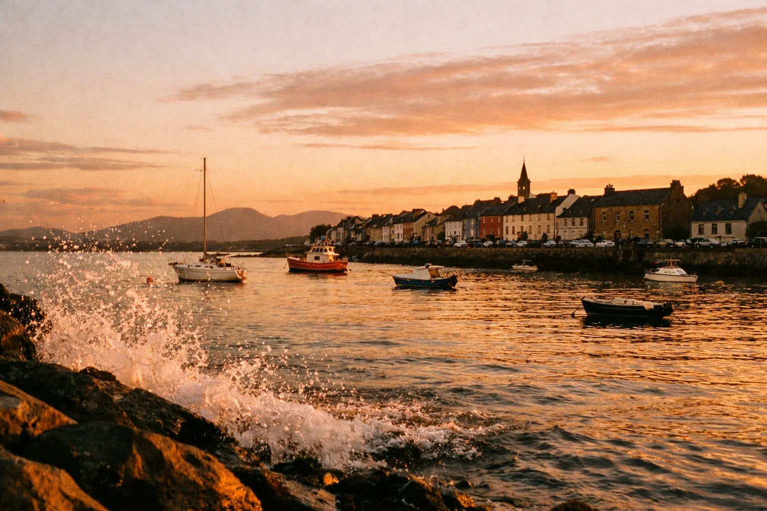 Boats float on the water near a row of colorful houses and a church tower in a coastal town at sunset.