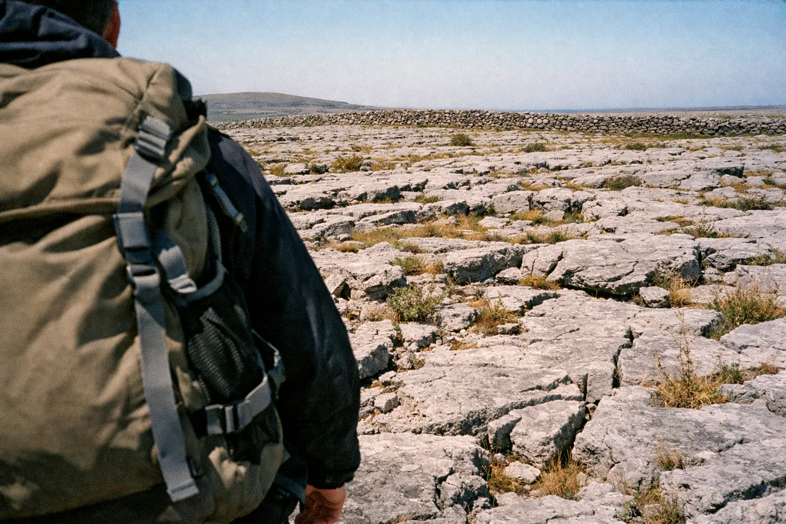 Person wearing a backpack walking on a rocky plain with sparse vegetation and a stone wall in the distance