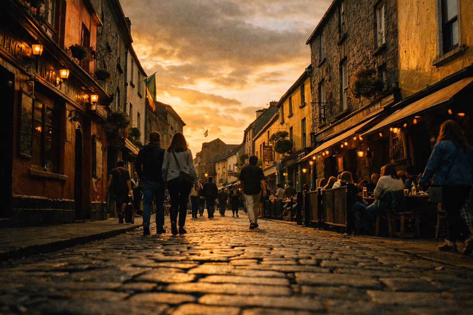 People walk along a lively cobblestone street lined with stone buildings and cafes at sunset in an Irish city.