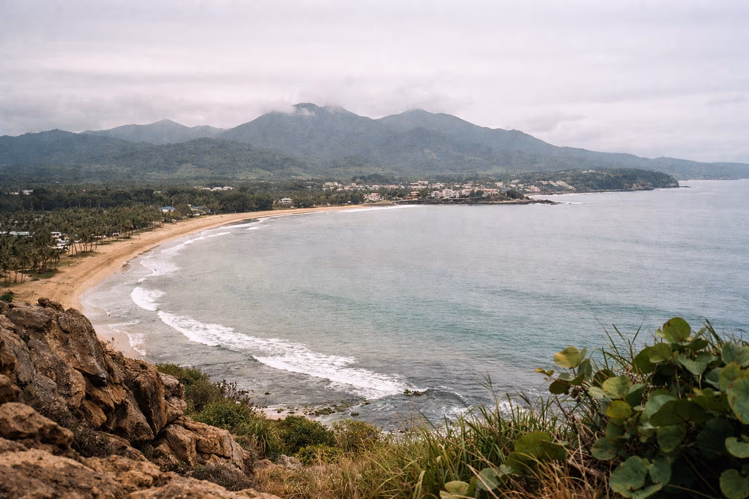 A wide sandy beach curves along the coastline with green mountains in the background in Luquillo, Puerto Rico.