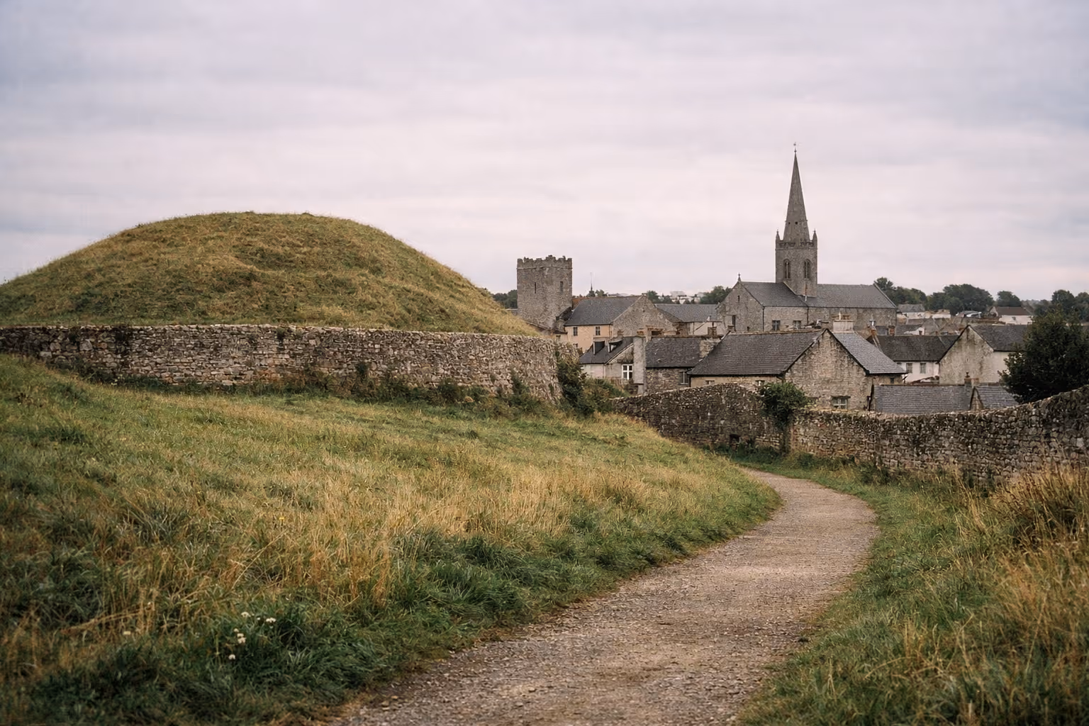 A grassy mound and stone wall stand before a village with a church and round tower in Navan, Ireland.