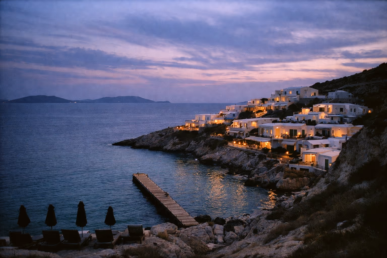 Coastal resort buildings with lit windows on a rocky hillside above the sea at dusk, with lounge chairs and umbrellas by the