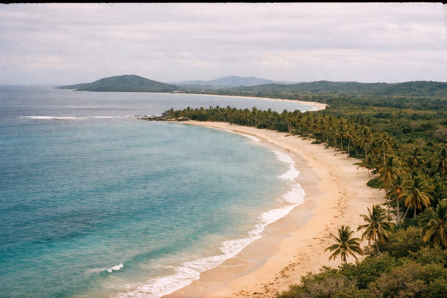 Curved sandy beach lined with palm trees next to turquoise water and lush green hills.