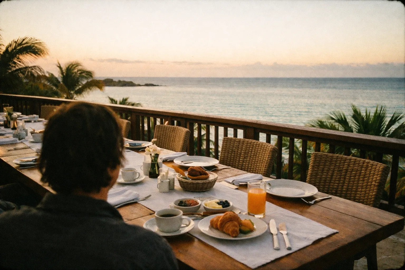 A person sits at a breakfast table on a terrace overlooking the ocean at sunrise.