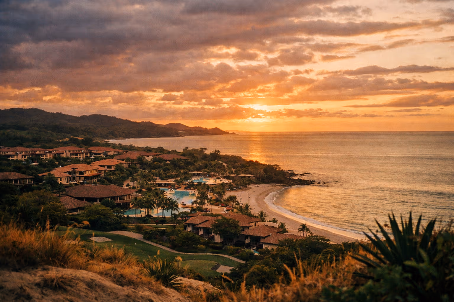 coastal resort with pools, red roof buildings, palm trees, and sandy beach at sunset