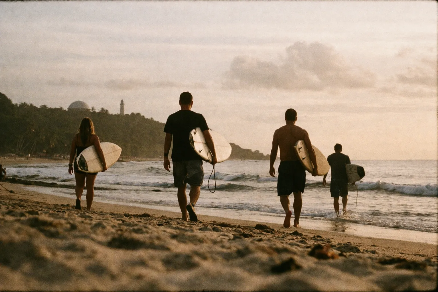 Four surfers walk along the sandy beach in Rincón, Puerto Rico at sunset carrying their surfboards.