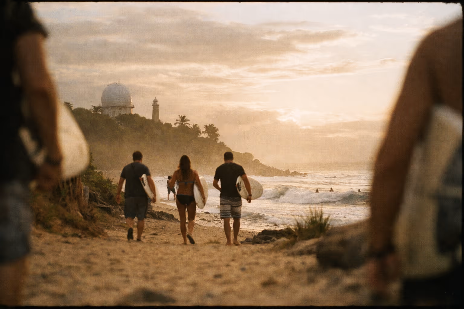 Surfers walk along a sandy beach in Rincón, Puerto Rico with a domed lighthouse visible on the bluff ahead.